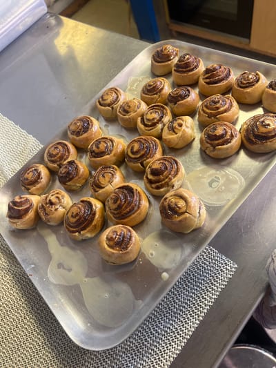 A high-angle, close-up photograph depicts a large, rectangular metallic baking tray, situated in the city of Ploemeur, France, containing approximately 30-35 freshly baked, round pastries identified as cinnamon rolls or similar spiral-shaped items. Each pastry features a golden-brown dough exterior with a visible dark brown, swirled filling, and is coated with a clear, glossy glaze. The glaze has partially dripped onto the reflective silver-toned metal tray surface, creating irregular puddles and streaks. The tray rests on a larger, equally reflective metallic countertop. To the upper left, a partially unrolled clear plastic film dispenser is visible. Sections of a white, open-weave mesh material are present on the metallic surface to the left and lower right of the tray. In the blurred background at the top right, a dark, rectangular appliance, possibly an oven, is partially visible.