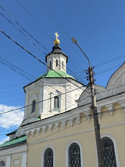 The image shows a view looking up at a church with a golden dome, situated against a bright blue sky. The church's architecture features a white facade with decorative elements and arched windows. A green roof is visible on one section of the building. A utility pole with numerous wires crisscrosses the frame, obscuring parts of the church and the sky. The wires are tangled and run diagonally across the image. The lighting suggests it is daytime, likely sunny, given the clear sky and shadows. The presence of the church with its dome and architectural style, combined with the provided location context of Yelets, Russia, indicates a religious building in that city. No people or specific events are depicted. The image is oriented sideways, with the church tower appearing to extend upwards from the bottom right of the frame.