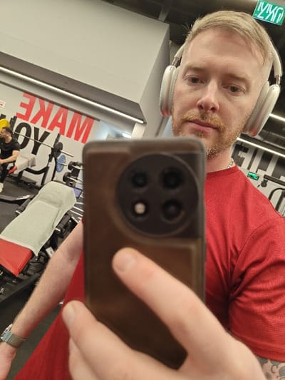 The image captures a selfie taken by a male individual in a mirror within an indoor gym setting in Israel, identified by the green Hebrew "יציאה" (EXIT) sign on the ceiling. The specific city of Nahariya is not visually confirmed.

The primary subject, positioned in the foreground, is a light-skinned male with short blonde hair, a reddish-blonde beard, and a mustache. He is wearing a red athletic short-sleeved shirt, a silver chain necklace, and light-colored over-ear headphones. A multi-colored beaded bracelet is visible on his right wrist. He holds a brown-cased smartphone with his left hand, and his right hand's fingers are partially visible. The smartphone's rear camera module, featuring a circular design with four lenses, is prominently out of focus in the foreground, indicating the act of photography.

In the background reflection, a second male individual is seated on a red and black weight bench. This individual has dark hair and a beard and is wearing a black t-shirt and black pants. A light grey towel is draped over the weight bench. Behind the seated individual, a barbell rack with a barbell and several circular weight plates (blue and black) is visible. A large graphic on the wall displays partial red and black text, reading "MAKE YOU." The gym features light grey walls, a white ceiling with linear fluorescent lighting, and a dark floor.