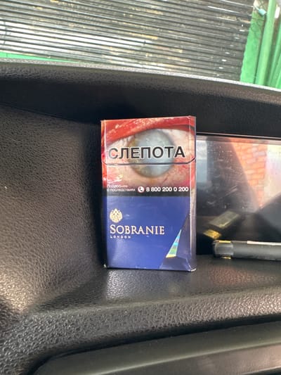 The image shows a pack of cigarettes with the brand name "SOBRANIE" printed on it, along with the word "LONDON". The pack also features an image of a human eye with red veins and the Russian word "СЛЕПОТА" which translates to "BLINDNESS". Below the eye image, there is text in Russian that says "Подробнее о последствиях" meaning "More about the consequences", and a phone number "8 800 200 0 200". The cigarette pack is placed on a textured black surface, possibly the dashboard or interior of a vehicle. In the background, there are horizontal metal blinds or shutters and a green curtain. The lighting suggests an indoor or covered environment. There are no people visible in the image. The location is identified as Nazran, Russia, but this specific detail cannot be visually confirmed from the image content alone.