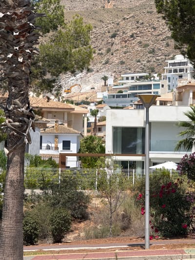The image depicts a hillside residential area in Urbanització Golf Badia, Spain, characterized by modern white multi-story houses and traditional terracotta-roofed villas nestled against a rocky, arid mountain slope. In the mid-ground, a large palm tree trunk is prominent on the left, with green pine trees to its right. An excavator is visible on the upper slope, suggesting ongoing construction or excavation work. The foreground features a neatly maintained garden with a white metal fence, various shrubs, and flowering plants, including pink oleander. A silver lamppost stands near the center of the frame. The overall impression is of a sun-drenched, Mediterranean landscape with a mix of architectural styles.