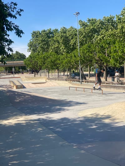 This media file captures an outdoor skatepark in Santa Perpètua de Mogoda, Spain, bathed in bright sunlight under a clear blue sky. The skatepark is constructed from concrete, featuring various ramps, ledges, and a quarter pipe, with distinct tree shadows falling across its surfaces.
The central subject is a young man, dressed in a dark t-shirt, dark shorts, white socks, and black shoes, actively skateboarding. He is positioned near a metal rail with several horizontal bars. In the background, another person, wearing a light-colored top and a cap, is also visible with a skateboard, standing near some trees and a fence. The skatepark is bordered by numerous leafy green trees, suggesting a park setting. A street light pole extends towards the top of the frame.
Notable details include the time of day, which appears to be morning or afternoon due to the long, sharp shadows cast by the trees, indicating a sunny and clear weather condition. On a concrete box/ledge in the skatepark, the word "Chipp'a" or similar is partially visible as text. The overall scene depicts a vibrant recreational activity in an urban outdoor environment.