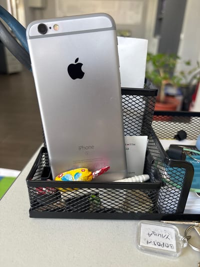 An iPhone 6S, silver, is positioned vertically within a black mesh desk organizer. The phone's screen faces away from the viewer, displaying the Apple logo and "iPhone S" inscription on its back. A yellow lollipop wrapped in colorful foil rests at the base of the phone, partially obscured by the organizer's front edge. Also visible within the organizer are various small items, including what appears to be a white tube and a small piece of paper with black text that reads "r. TTH" and "ua." In front of the organizer, a clear plastic keychain with handwritten text in what appears to be Cyrillic script and a silver keyring are visible on a light-colored surface, likely a desk. The background is blurred, suggesting an indoor setting with a plant visible in the upper right quadrant. The lighting is bright, casting subtle shadows. The scene is located in Pereiaslavske, Ukraine.