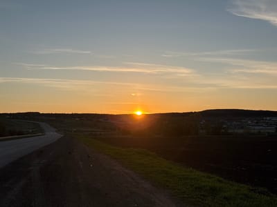 The image depicts a sunset view in what appears to be a rural area, possibly near Bagrash-Bigra, Russia. A road stretches into the distance, curving slightly to the left. The sun is setting directly ahead, with a golden glow obscuring some of the details on the horizon. The landscape includes fields, some of which are ploughed, and patches of trees. Buildings or houses are faintly visible in the distance, suggesting a village or small town nearby. In the foreground, the edge of a dirt road is marked with green grass. There are some dark-colored small birds on the asphalt. The sky is a gradient of light blue and orange, with thin wisps of clouds near the horizon.