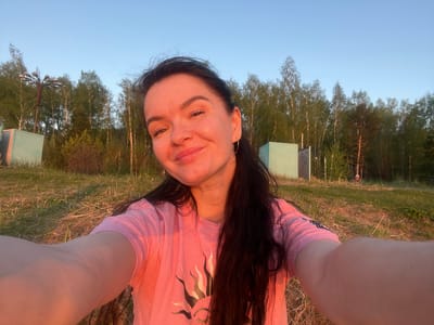 The image shows a woman taking a selfie in Borovoye Matyushino, Russia. She is wearing a pink t-shirt with a sun design. The background includes trees, grass, and what appears to be street lighting or signs. The sky is visible above the trees.