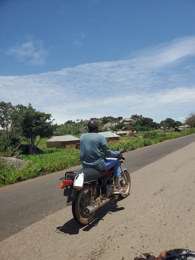 A person riding a motorcycle travels on a paved road through a rural area, possibly Panyam, Nigeria. The rider, seen from behind, wears a blue long-sleeved shirt and blue jeans. Their feet rest on the motorcycle's footpegs. The motorcycle is red and has a black seat and a white bottle attached to the rear. A shadow of the motorcycle and rider is cast on the road. The landscape features lush green vegetation, scattered trees, and rocky hills in the background. Several small structures with corrugated metal roofs are visible amidst the greenery, indicating a settlement. The sky is a bright blue with scattered white clouds.