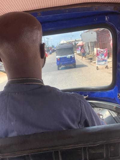The image presents a street scene in Anantigha, Nigeria, observed from inside a vehicle, looking forward over the driver's left shoulder. The driver, an adult male with a bald or closely shaved head, wears a dark blue collared shirt featuring a striped pattern on the collar. The interior of the viewing vehicle includes a blue frame and a light-colored, patterned ceiling fabric. Ahead, a blue and black three-wheeled vehicle (tricycle/Keke Napep) with "GENERAL GOODS" printed on its rear and a partially visible license plate is proceeding on a paved road. To the right of the road, a dirt shoulder and a drainage ditch are visible, alongside a person in a red shirt walking while carrying an object. A prominent mobile A-frame sign, predominantly pink and white, advertises "POS" services. The street is lined with various single-story structures, consistent with commercial buildings, under a partly cloudy sky.