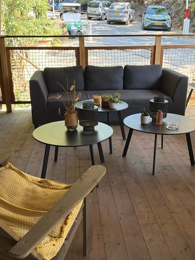 A group of cars and a golf cart are parked on a paved area adjacent to a wooden deck. The deck features a dark gray sofa, three black circular coffee tables, and a wooden chair draped with a yellow blanket. On the coffee tables are decorative items such as lanterns, small plants, a vase, and an ashtray. The scene appears to be an outdoor seating area. The provided information indicates the location is Castelnuovo dei Sabbioni, Italy.
