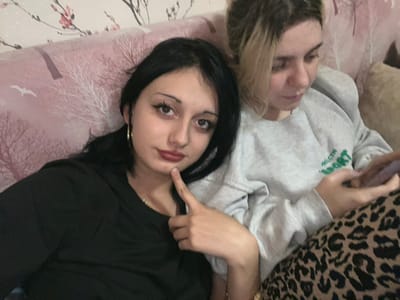 Two females are depicted indoors, seated on a sofa in Lanchkhuti, Georgia. The sofa's backrest is upholstered in a pinkish fabric patterned with white-branched trees and small birds. A plain beige cushion is partially visible on the right.
The female on the left has long, dark hair and is wearing a black long-sleeved top and gold-colored hoop earrings. Her face features visible dark eyeliner and reddish lipstick. Her right index finger is pressed against her chin, and she is looking directly at the viewer with a neutral expression.
The female on the right has lighter, possibly blonde or light brown, hair and is wearing a light grey long-sleeved sweatshirt with a partial green "MILAN SPORT" logo on the front. Her lower body is covered by leopard-print fabric, likely pants. She is looking down, engaged with a rectangular electronic device, presumably a smartphone, held in both hands. She appears to be interacting with the device.