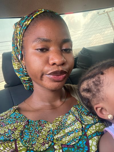 A woman with brown skin and a patterned headscarf poses with a baby whose head rests on her shoulder. The baby has an earring and is wearing a patterned top with embellishments. The woman is also wearing a top with a similar green and blue pattern. The photo appears to be taken inside a car, as a window and car seat are visible in the background.