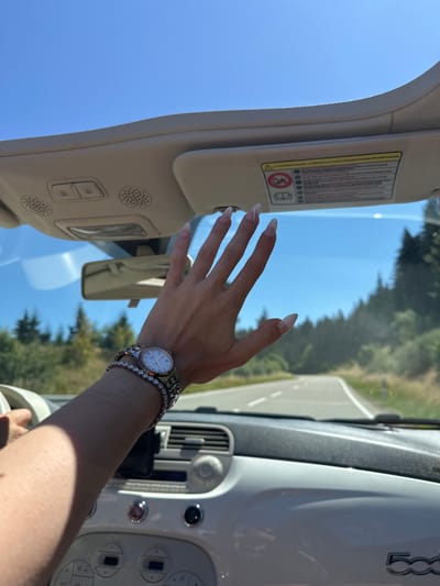 The image captures a first-person perspective from inside a convertible, likely a Fiat 500, as evidenced by the "500" emblem visible on the dashboard. The car is on a paved road that curves to the right, surrounded by a lush, green landscape with numerous conifer trees. The sky is a clear, bright blue. In the foreground, a person's right arm and hand are visible. The hand is raised, with fingers extended, and has long, manicured nails with a French tip design. The wrist is adorned with a silver watch with a white face and a delicate bracelet, along with a second sparkly bracelet underneath. The dashboard is white and features various buttons and controls. The sun visor above the windshield has a yellow warning sticker with text in multiple languages. The scene suggests a scenic drive on a sunny day. The car's location is Furtwangen im Schwarzwald, Germany.