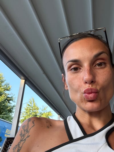 This is a close-up, selfie-style photograph of a woman in Razliv, Bulgaria. She is outdoors, seated under a white slatted awning. The woman has brown hair, brown eyes, and light skin with freckles. She is wearing a white tank top with black trim and has a tattoo of a floral design on her left shoulder and collarbone. She is puckering her lips, and a pair of black-rimmed glasses are perched on top of her head. In the background, through a window or opening, are green trees and a bright blue sky. A blue truck with yellow lettering is partially visible on the left side of the frame.