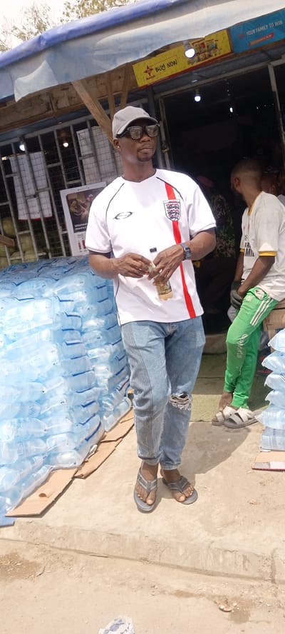 The image shows a man standing in front of what appears to be a retail establishment or market stall. The location can be identified as the city of Gayawa, Nigeria.

The man is wearing a white t-shirt with a red stripe on the side and an England football crest on the chest, light blue jeans with a rip at the knee, a gray baseball cap, and dark sunglasses. He is holding a small bottle in his hands.

To his left is a large stack of bottled water. In the background, there's another person wearing a white t-shirt and green pants, seated, with more stacks of bottled water nearby. There are signs above the stall, some featuring advertising and brand names. The stall appears to be constructed with a corrugated metal roof. The ground is a light-colored concrete or paved surface.