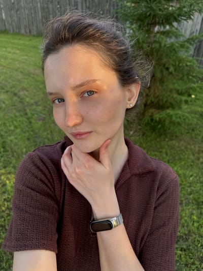 A young woman with fair skin, light brown hair, and blue eyes is outdoors, possibly in a suburban backyard in Burdyata, Russia. She is wearing a dark maroon, short-sleeved, collared knitted shirt. Her right hand is raised, with her fingers resting on her chin and jawline. She is also wearing a silver-banded fitness tracker on her left wrist. The background consists of a wooden fence, green grass, and the foliage of a coniferous tree. The lighting suggests it is late afternoon or early evening, with the sun casting a warm glow on her face.