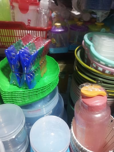 The image shows a collection of household plastic items stacked together. A stack of green leaf-shaped plates topped with packaged clothespins are prominent. Adjacent are stacks of colorful buckets and basins, with pink and yellow accents. In the background, there are containers and dispensers in red and transparent plastics. The foreground includes clear plastic containers and a pink water bottle with an orange lid. The overall impression is a market or storage area displaying various plastic goods.