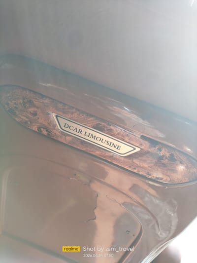 The image shows a close-up view of a car's interior, specifically a wooden trim piece with the text "DCAR LIMOUSINE" embossed on a metal plate. The wood grain pattern is intricate and appears to be a veneer. The surrounding interior surfaces are a glossy brown color, showing some signs of wear and minor cracks. In the lower right corner, a watermark indicates "realme Shot by zsm_travel 2026.06.24 07:50". The location is not discernable from the image itself, and there are no people, actions, or interactions depicted. The environment is limited to the interior of a vehicle.