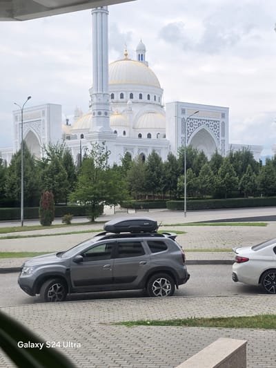 The image captures a view of the Akhmad Kadyrov Mosque in the city of Shali, Russia, on an overcast day. In the foreground, a grey SUV with a roof cargo box is parked on the street. To the right of the SUV, the rear of a white sedan is visible. The mosque, a prominent landmark, features white architecture with golden domes and minarets, surrounded by lush green trees. A sidewalk with a paved pattern is visible in the lower part of the image, and a white stone ledge is present in the bottom right corner. The sky is predominantly cloudy.