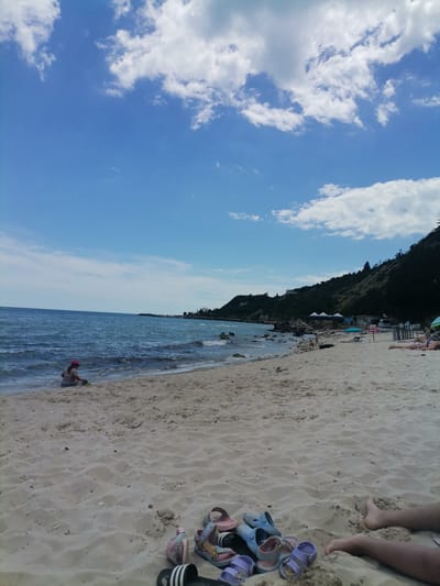 A wide, sandy beach is pictured under a bright blue sky with scattered white clouds. In the foreground, a collection of colorful children's sandals and flip-flops are laid out on the sand. The middle ground shows the gentle waves of the sea lapping at the shore. A young child, wearing a pink hat and a patterned swimsuit, is playing in the shallow water near the edge of the beach. Further back, a dark, tree-covered hill rises from the coastline, with a few buildings and tents visible at its base. A few other beachgoers are present, some lying on towels and others standing near the water. The scene depicts a relaxed day at the beach in the city of Osenovo, Bulgaria.