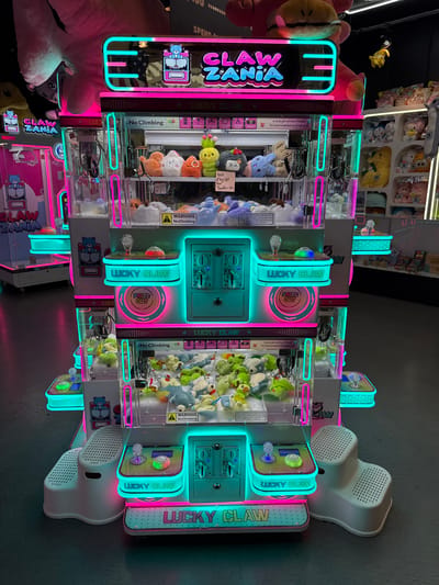 The image depicts a brightly lit arcade, likely a "Claw Zania" themed establishment in El Paso, United States of America. The central focus is a large, multi-tiered claw machine filled with plush toys. The machine features prominent neon pink and teal lighting, along with digital displays and control panels. Several other claw machines are visible in the background, some also displaying plush toys. The floor appears to be a dark, polished surface, reflecting the vibrant lights of the arcade. There are no people visible in the image, and the scene is static, showcasing the arcade machines in operation.