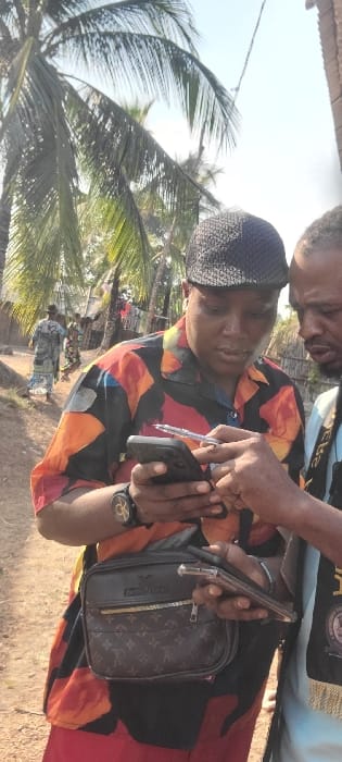 The image captures an outdoor scene in Alayi, Nigeria, featuring two men interacting while focused on a smartphone. The primary subject, positioned on the left, wears a black patterned flat cap, a short-sleeved collared shirt with a vibrant abstract pattern (red, orange, black, yellow), and red trousers. He holds a black smartphone with a clear case in his left hand and points a clear-barreled pen towards the screen with his right hand. A black wristband watch with a gold circular emblem is visible on his left wrist, and a brown shoulder bag with a patterned front is slung across his body. The second man, partially visible on the right, wears a light blue garment with a black and gold fringed sash. His left hand is near the first man's phone, and he holds a second smartphone with a clear case in his right hand. Both individuals' gazes are directed at the screen of the phone held by the first man. The background reveals several tall palm trees under a bright sky, unpaved ground, and distant rudimentary structures, including one with clothes hanging on a line. Two additional figures are visible walking in the far left background, one wearing a patterned long dress.
