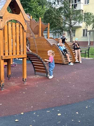 In Viazniky, Russia, a playground scene unfolds with four people interacting on a wooden play structure. The structure features curved wooden elements and a slide. Three children and one adult are visible. A young girl in a pink shirt and blue overalls is facing away from the camera, with her hands on the play structure, appearing to be walking or climbing. Two boys are lying down on the wooden structure. One boy, wearing a black and white striped shirt and black shorts with white accents, is lying on his back. Next to him, another boy in a grey shirt and black shorts is also lying down. An adult in a black top and light-colored pants is lying down further up the structure, looking at their phone. The ground is a rubberized surface, with patches of red and blue. Fallen leaves are scattered across the ground. The time appears to be daytime, with ambient light suggesting it is not midday. The overall mood seems relaxed and casual, with the individuals enjoying the playground. A building and trees are visible in the background. No visible text is present.