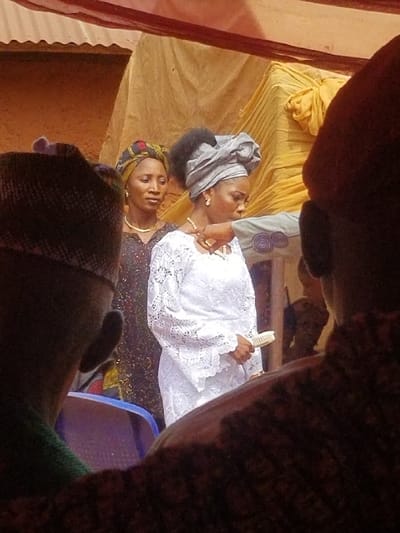 The image captures an outdoor or semi-outdoor gathering in Ochadam, Nigeria, observed from the perspective of two individuals partially visible in the foreground. Overhead, a reddish-brown fabric canopy transitions into a yellow fabric canopy.
In the midground, three individuals are prominent. On the left, a woman with medium-dark complexion wears a multi-colored headwrap (gele) and a dark, patterned dress with a gold-colored necklace and earrings, looking slightly to the right. Central to the frame is another woman with medium-dark complexion, wearing an elaborate light-grey headwrap, a white long-sleeved lace dress with a distinctive pattern, a gold-colored necklace, earrings, and a bracelet. She holds a small, light-colored, rectangular object in her right hand and is looking downwards and to her right. An arm from the right side of the frame, clad in a light-colored garment with a spiral pattern on the sleeve, extends towards and is positioned near the right shoulder of the central woman.
The background features an earth-toned wall structure, possibly unpainted concrete or mud, and a larger object draped in yellow fabric. A blue plastic chair is partially visible in the lower-left midground. The foreground shows the back of a dark cap and a green garment on the left, and a dark, patterned garment on the right, belonging to observers.