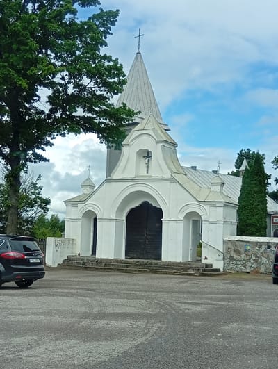 The image shows the entrance of a white church in Šaukėnai, Lithuania. A dark colored SUV is parked to the left of the frame. The church has a tall, pointed steeple with a cross at the very top. The facade is white with arched doorways and decorative elements. A large, dark wooden door is visible behind the central archway. To the right of the church entrance, there is a low stone wall with a yellow scallop shell symbol, indicating a pilgrimage route. The ground in front of the church is paved with asphalt. Tall trees and a cloudy sky form the background.