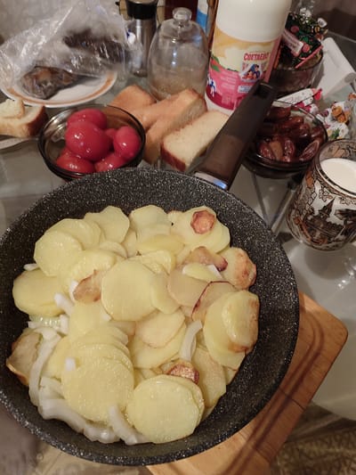 The image shows a close-up of a meal laid out on a table, likely for lunch or dinner. The primary focus is a frying pan filled with sliced potatoes and onions, some of which appear to be fried. In the background, other food items are visible, including a bowl of cherry tomatoes, several slices of bread, a container of dates, and a glass or mug filled with a white liquid, possibly milk. There is also a plastic-wrapped item that seems to contain some kind of dried fish or meat. A bottle with a label featuring the word "СВЕТАЕВО" and an illustration of children is prominent. The setting appears to be indoors, with a table surface and what looks like a tiled floor visible. The lighting suggests it might be daytime, but the specific time of day is not definitively indicated. There are no people visible in the image. The overall impression is of a home-cooked meal being prepared or served. The location context is Abinsk, Russia, which aligns with the Cyrillic text on the bottle.