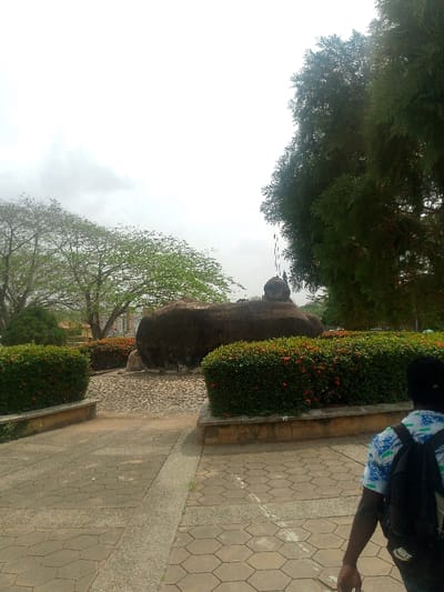 The image displays an outdoor scene in Adzho, Nigeria, under an overcast sky. In the immediate foreground, the back of an individual is visible on the right, wearing a patterned light blue and white short-sleeved shirt and a black backpack, walking away from the camera. The ground is paved with hexagonal and rectangular interlocking concrete pavers, bisected by a lighter concrete strip.

Midground features a central open area flanked by two raised planter beds constructed of concrete and stone, densely planted with green bushes exhibiting small red flowers. Beyond these planters, a prominent, dark, irregularly shaped rock formation occupies the center, appearing as several connected boulders, with a smaller, rounded rock atop the right-hand portion. The ground immediately surrounding the rock formation is paved with irregular cobblestone-like stones.

The background consists of numerous trees with varying shades of green foliage; a large, dense tree with darker green leaves is visible on the far right, while trees with lighter green leaves and more visible branches are on the left. Faint outlines of distant structures are discernible through the trees on the left. A thin, dark vertical object, possibly a pole, extends upwards above the right side of the rock formation.