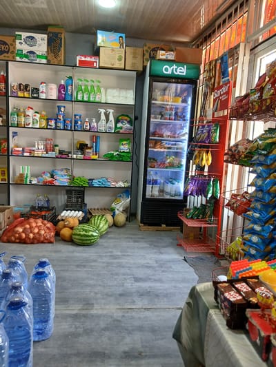 This is a medium shot, eye-level, indoor photograph of a small convenience store, likely located in Denov, Uzbekistan, given the product packaging and general store setup. The scene is brightly lit, possibly by overhead fluorescent lights visible at the top of the frame, and natural light coming from a doorway or window on the right.
The store is stocked with a variety of merchandise displayed on white shelves and in a glass-fronted refrigerator. The shelves are filled with household cleaning products, toiletries, packaged foods like biscuits and snacks, and bottled beverages. Some items are in their original packaging, while others appear to be individually placed.
On the floor, in the foreground and midground, are various food items and large water bottles. A large net bag filled with onions is prominent on the left. Next to it are several watermelons, pumpkins, and possibly other fruits and vegetables. Several large, clear plastic bottles of water are lined up on the floor in the bottom left quadrant of the image.
To the right, a red metal display rack is filled with bags of chips and other snacks. A glass-fronted refrigerator with an illuminated "artel" logo on top is situated on the left side of the store. Inside the refrigerator, shelves display various food and drink items, including bottles and containers.
A table covered with a patterned cloth is visible on the far right, displaying more packaged snacks and candies. The overall impression is of a well-stocked, albeit somewhat cluttered, local grocery or convenience store. There are no people visible in the photograph. The flooring appears to be a gray, possibly concrete or tiled, surface. The ceiling is made of a white, ribbed material.