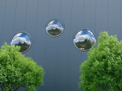 The image displays the exterior of a modern building in Peetri alevik, Estonia, characterized by a dark grey, paneled facade. Three circular, convex windows are evenly spaced on the wall, each reflecting a landscape of trees and a sky filled with dramatic white clouds. In the foreground, two lush green trees with delicate foliage flank the building, partially obscuring the lower portions of the windows and the facade. The lighting suggests it is daytime with overcast skies, casting a soft light on the scene. There are no people or any discernible actions taking place.