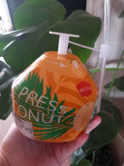 An orange, stylized coconut-shaped beverage container, labeled "PRESS COCONUT" in white text with green palm frond graphics, is held by a light-skinned human hand. The container features a white plastic dispenser and an attached clear plastic straw, secured by a white plastic clip. A red octagonal label reading "HOW TO DRINK" is visible upside down on the container. In the background, a large, dark green leaf of a houseplant, consistent with a Monstera deliciosa, is visible, with a light-colored curtain or wall and a bright window behind it, indicating an indoor setting. This scene is located in the City of Drama, Greece.