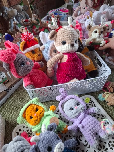 The image displays a vibrant collection of numerous handmade crocheted plush toys, primarily animals, artfully arranged within and around white decorative plastic baskets. The setting appears to be a market stall or craft fair, with the items resting on a green, textured woven surface.
**Subjects:** The central subjects are a wide array of soft, colorful crocheted animals. These include multiple deer or reindeer figures, some in white with brown antlers, others in brown, and some adorned with red and white hats. Various bunnies are also prominent, appearing in shades of purple, pink, and grey, some with winking eyes or little noses. Other animal-like figures are seen in bright orange, green, and yellow. A human hand is visible in the top left corner, seemingly interacting with the display.
**Objects:** The toys are contained within and spilling out of white plastic baskets, characterized by intricate, cut-out patterns. The items are showcased on a distinct green, textured mat or fabric. Small wooden and metallic decorative elements, including miniature reindeer ornaments, are visible in the background, hinting at a festive or holiday theme.
**Activity:** The scene captures the presentation or sale of these handcrafted items. The presence of the human hand suggests someone is either arranging the display, browsing the merchandise, or making a selection.
**Notable details:** The crocheted toys are crafted from plush, likely velvet or chenille, yarn, giving them a soft, inviting texture. They feature charming details such as embroidered eyes, rosy cheeks, small bows, scarves, and hats. The color scheme is diverse and cheerful, incorporating pastels like lavender and baby pink, alongside bolder hues of fuchsia, orange, green, and red. The overall impression is one of handcrafted warmth and whimsy.
**Visible text:**
* A yellow price tag on one of the white baskets shows "1.200" and below it, partially visible Cyrillic text that translates to "toy" (игрушка).
* A small white tag attached to a green-hooded orange toy has the English phrase "with love" printed on it.
* Near the bottom right, on a small white card, the word "MECLUB" is visible.
The context of Ludorvai, Russia, aligns with the Cyrillic text observed on the price tag, indicating a local market or craft event.