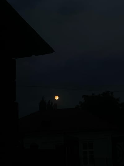 An orange moon is visible in the dark night sky above rooftops and silhouetted trees in Myrza-Ake, Kyrgyzstan. A dark, angular structure, possibly a building or eaves, juts into the frame from the left. Power lines are strung across the upper portion of the sky. The scene suggests a quiet, possibly residential area at dusk or after nightfall.