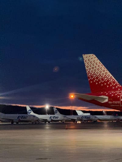 The image shows a row of UTair airplanes parked on an airport tarmac at dusk, with a red and white tail fin of a separate airplane in the foreground. The location is most likely Емельяново аэропорт, Russia. The sky is dark blue, transitioning to orange near the horizon where the sun has set. The airport tarmac is illuminated by overhead lights, reflecting off the wet surface. The UTair airplanes are white with blue lettering. The red and white tail fin has a geometric pattern.