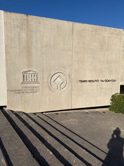 The image depicts the exterior of a building with embossed text and logos. On the left, the UNESCO logo and the full name "United Nations Educational, Scientific and Cultural Organization" are visible. To the right of this, the UNESCO World Heritage emblem is embossed. Further to the right, the text "TEMPLJI NEOLITICI TAL-ĠGANTIJA" is inscribed. The building's facade is a textured, light-colored stone, with a distinct vertical seam running down the center. In front of the building, a dark-colored paved area with diagonal shadow lines from an unseen structure or object is present. A shadow of a person taking the photograph is visible in the lower right corner. A green hedge is located on the far right edge of the image. The sky above is a clear, vibrant blue. The building is identified as the "Ġgantija Temples" located in Xagħra, Malta.