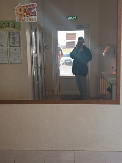 The image shows a reflection in a mirror. The reflection depicts a person taking a photo of themselves in front of a doorway. Outside the doorway is a street scene with cars and people. Inside the doorway is a room with windows, documents on the wall, and a poster that reads "9 MAY 1941-1945", the Russian Victory Day. "GORY" is visible in Russian. The location is likely in Gory, Russia.