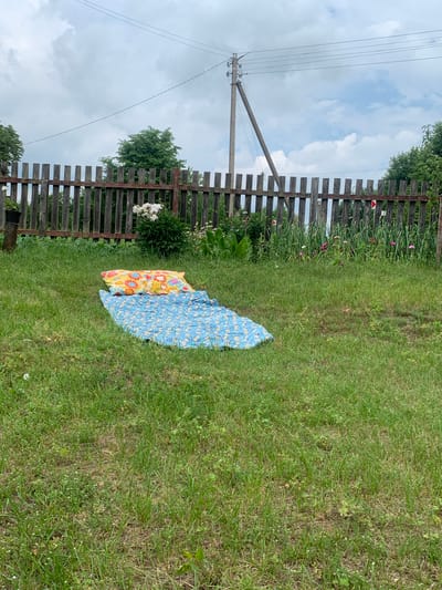 A picnic blanket is laid out on a grassy lawn in a rural setting. The blanket is blue with a floral pattern and lies next to a yellow blanket with a floral pattern. A wooden fence runs along the left side of the image, separating the lawn from a garden with blooming pink and white flowers. Beyond the fence, a telephone pole with wires stands against a cloudy sky. The scene appears to be set in the afternoon on a partly cloudy day, suggesting a casual outdoor activity like a picnic or a place to relax. The location is identified as Орлеўскі сельскі Савет, Belarus. There is no visible text in the image.