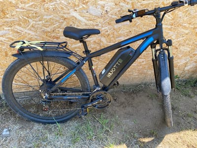 A black DUOTTS electric bicycle, adorned with blue geometric accents on its frame, is parked outdoors against a textured wall made of Oriented Strand Board (OSB). The bicycle features a large black battery pack with the DUOTTS logo in a green camouflage pattern mounted on the downtube. A rear hub motor is integrated into the black rear wheel, which is fitted with a knobby Chao Yang tire. The drivetrain includes a Shimano rear derailleur and multi-speed chainrings. Front suspension forks with silver stanchions are visible above the front wheel, also equipped with a knobby tire and a black fender. A black cargo rack is present over the rear wheel, secured with a yellow, possibly fabric, strap. The handlebars are equipped with black grips, brake levers, shifters, and a small digital display unit to the left of the stem. The ground beneath the bicycle is uneven, consisting of light brown dirt mixed with sparse green grass and dried vegetation. The scene is brightly illuminated by natural light, casting subtle shadows. No individuals are present in the image. There is no visual information within the image to confirm the specific location as the city of Belo Pole, Bulgaria.