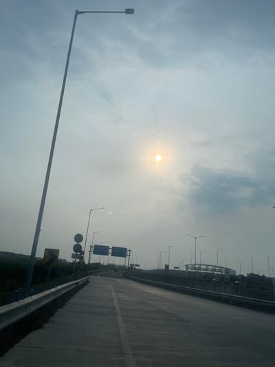 The image depicts a view of a highway on a hazy day, with the sun partially obscured by clouds and casting a bright glow. A tall street light stands prominently on the left side of the frame. The highway curves to the right, with multiple lanes visible. Road signs, including directional signs and a warning sign with a yellow chevron, are present. Several other street lights line the highway. In the background, there appears to be some construction or an elevated structure. The environment suggests an urban or suburban setting in Salembaran Jati, Indonesia, given the presence of infrastructure like a highway and street lights. No people are clearly visible in the frame.