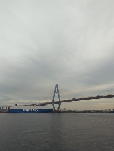 The image shows a car carrier ship named "FUJITRANS" docked in Nagoya, Japan, likely in the Port of Nagoya. The ship is large and blue and white, with "FUJITRANS" written in large white letters on its side. In the background, a large suspension bridge spans the water, with cars visible on its roadway. The sky is overcast with gray clouds. The water is dark and calm. The overall scene suggests a bustling port environment with significant industrial infrastructure.