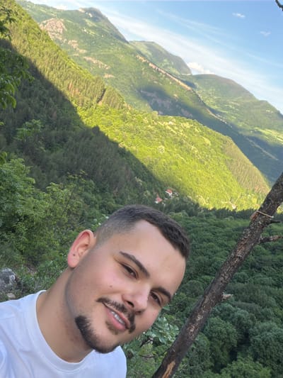 A selfie of a young man with light skin, short dark hair, and a dark mustache and beard, wearing a white T-shirt. He is positioned to the right of the frame. The background is a mountainous landscape densely covered with green trees, typical of a wooded area. A single, charred branch diagonally crosses the upper right portion of the frame, partially obscuring the background. The left side of the frame showcases a valley with varying shades of green vegetation. The sky is visible in the upper left corner, revealing a clear blue expanse with a few scattered clouds. Based on location information, the image likely depicts a setting in or near Zasele, Bulgaria, given the area's geography and environmental attributes.