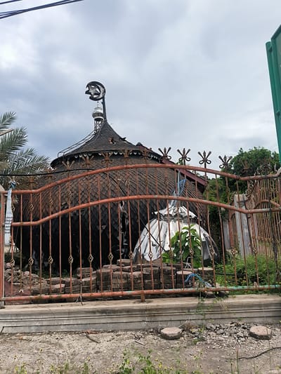 The image depicts a unique, circular building, likely a mosque or prayer hall, situated in Kuta Pungwa, Indonesia, under an overcast sky during daytime. The building features a dark, rounded roof covered in a pattern of small, overlapping shingles, giving it a scaled appearance. A tall, slender structure resembling a minaret rises from the center of the roof, topped with a prominent crescent moon and star finial, a symbol of Islam.
In the foreground, a heavily rusted, ornate metal fence spans horizontally across the frame, partially obscuring the lower section of the building. The fence has decorative spiral and star-shaped elements. Behind this fence, to the right of the main building, is a smaller, shiny, reflective dome-like structure, possibly a planter cover, with green plants growing inside. Piles of bricks or pavers are visible behind the fence.
To the left, palm fronds are visible. The ground in the immediate foreground consists of concrete and dirt with some weeds and scattered debris. Utility lines stretch across the upper left portion of the sky, and a partial green signpost is visible on the far right. A tattered white banner or sign hangs from the fence on the left, displaying partial Indonesian text including "...ASIL," "Laborat...", "budaya," "Musholla," "lapang...", and "Menda...". The scene conveys a rustic and somewhat weathered atmosphere.