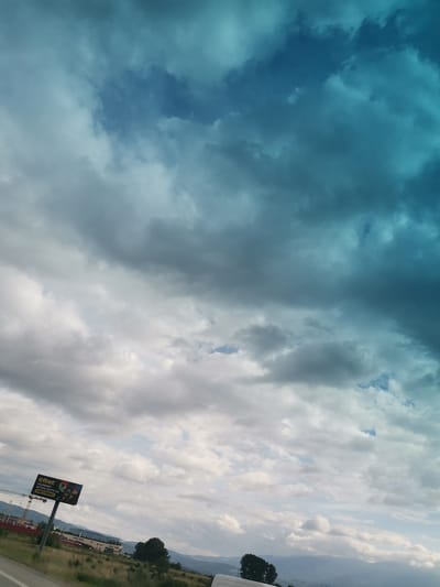 The image depicts a vast, cloudy sky with a turquoise hue transitioning to gray and white. In the foreground, a billboard stands to the left, advertising "efbet" with Bulgarian text. Below the sky, a landscape is visible, featuring a road, dry grass, trees, buildings, mountains in the distance, and construction cranes. A white van is partially visible at the bottom right. The scene appears to be captured from a moving vehicle, likely on a highway in or near Krivina, Bulgaria.