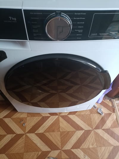 The image shows a close-up of a white washing machine. The washing machine has a control panel with a dial and several program options. The options visible include "Моя программа" (My program), "Синтетика" (Synthetics), "Хлопок" (Cotton), "Смешанная стирка" (Mixed wash), "Детская одежда" (Baby clothes), "Деликатная 20°C" (Delicate 20°C), and "Удаление шерсти" (Wool removal). On the right side of the panel, there are options for "Быстрая 15 мин" (Quick 15 min) and "Быстрая 59 мин" (Quick 59 min), "Эко 40-60" (Eco 40-60), "Шерсть" (Wool), "Постельное белье" (Bed linen), "Джинсы" (Jeans), "Отжим" (Spin), "Температура" (Temperature), and "Отложенный старт" (Delayed start). The machine also has a large, black, circular door with a patterned glass insert. The washing machine is positioned on a floor with a geometric wood-patterned tile. The background of the image is out of focus but appears to be a wall or room interior. The scene does not contain any indications of the city of Шипуново, Russia.