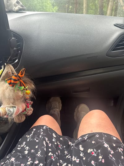 The image shows the interior of a car, looking forward from the passenger seat. The dashboard is visible, as is the leg of the person taking the photo, wearing a black floral dress and light-colored sneakers. To the left of the frame, a stuffed gnome-like figure with a St. George's ribbon and a small orange decoration is hanging from the dashboard, with rolled-up banknotes tucked into its fabric. The view through the windshield shows trees and greenery, suggesting an outdoor or roadside location. The specific location in Stepanovo, Russia is not discernible from the image itself.