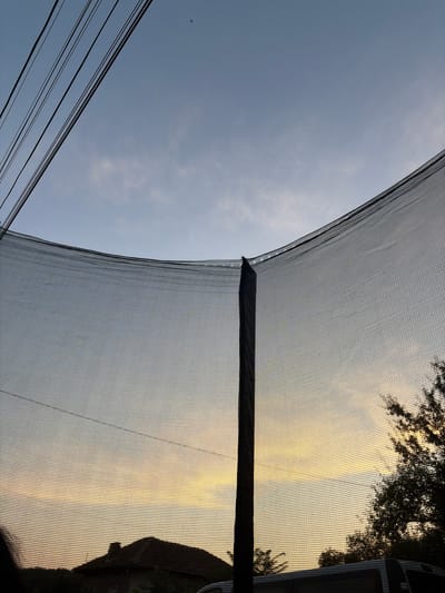 The image shows a view of the sky through a trampoline net at sunset. Several power lines are visible in the upper left corner against a pale blue sky. The setting sun casts a warm yellow and orange glow across the horizon, partially obscured by the net. In the lower portion of the image, the silhouette of a roof of a building and trees are visible. The image appears to have been taken from within a residential area in Karamanovo, Bulgaria, as suggested by the presence of the house and trees.