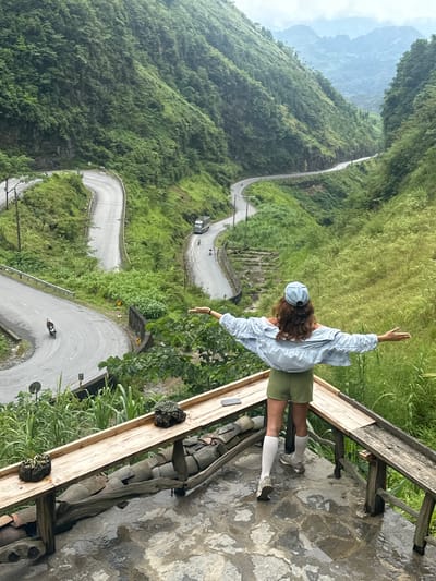 A bird's eye view shows a person standing on a stone patio with a wooden railing in Phố Bảng Commune, Vietnam. The person, presumably a woman with curly brown hair, wears a light blue hat, light blue long-sleeved button-down shirt, green shorts, white socks, and grey sneakers. She is standing with her arms outstretched, facing away from the camera. A long, winding road is visible below, cutting through a lush green landscape. Several vehicles are on the road, including a bus and motorbikes. Mountains are visible in the distance. The patio is paved with stone and has a rustic wooden railing. The lighting suggests it is a bright day, possibly overcast. The scene conveys a sense of freedom and awe in a mountainous landscape.
