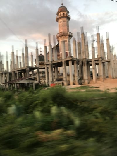 A horizontal view of a cityscape, shot outdoors during daylight. On the left is a tall, tan, and beige minaret with a bulbous dome. Adjacent to the minaret are numerous concrete pillars with exposed rebar, suggesting an ongoing construction site. The background sky is cloudy. To the right, blurred green foliage indicates the presence of vegetation. The location is Keudealue Ie Puteh, Indonesia.