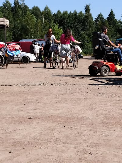 In Irshansk, Ukraine, on a sunny day, an outdoor market scene unfolds. The foreground is dominated by a dusty, unpaved ground. In the mid-ground, two women are walking with bicycles. One woman, with long brown hair, wears a plaid shirt and shorts, with a backpack. The other woman, with long dark curly hair, wears a pink shirt and denim shorts. To the right, a man sits in a red mobility scooter, wearing a gray shirt, blue jeans, and a dark baseball cap. He is looking to his right, and a yellow bag is attached to the scooter. Behind the women, a white car is parked next to a red van with its rear doors open. Further back, a tent structure with bundled textiles is visible. The background is filled with a dense line of green trees under a clear blue sky.